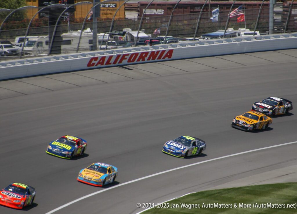 Several race cars speed around a curve on a NASCAR track at California Speedway, with a train visible in the background.