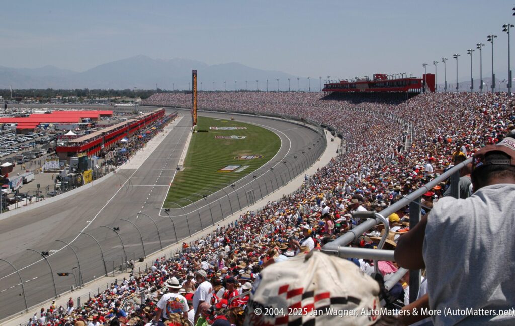 A large crowd fills the grandstands at an outdoor racetrack during a car race, with the track curving to the left and pit lane visible on the infield.