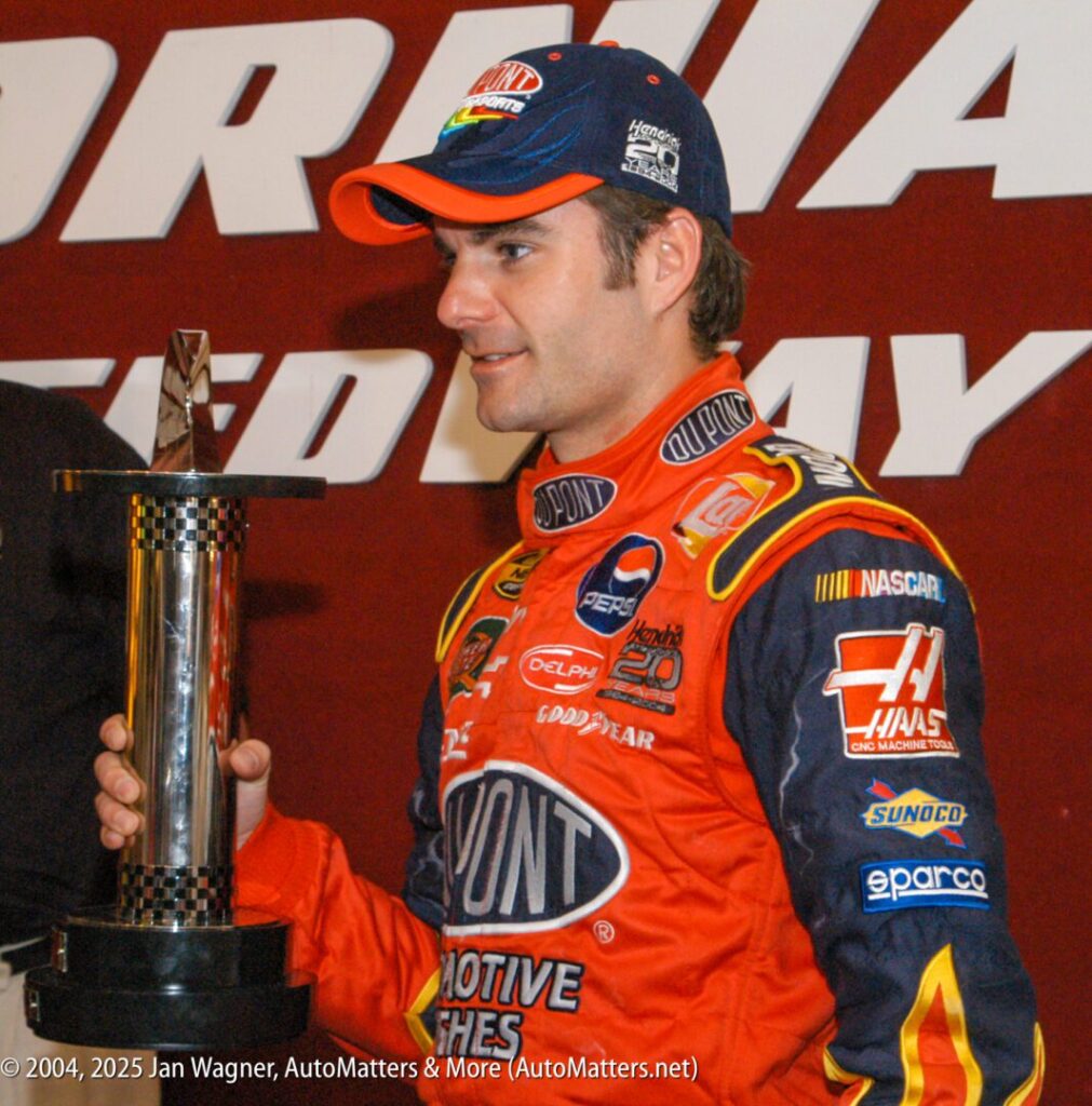 A race car driver in a red and blue firesuit holds a tall trophy in front of a sign that reads "Speedway.