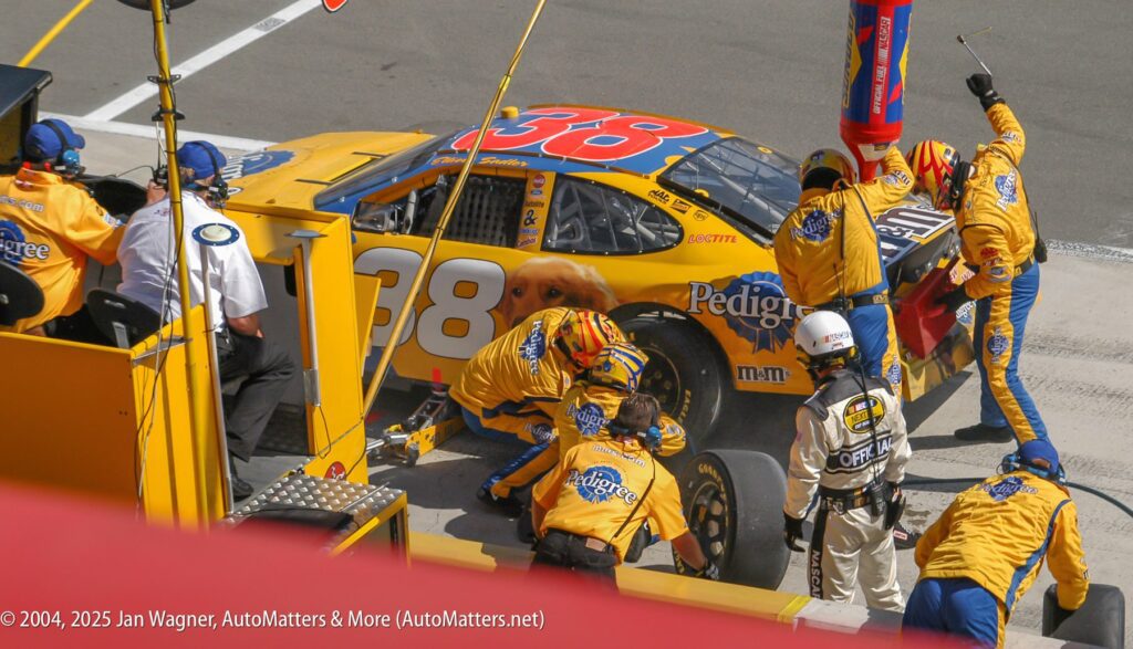 A NASCAR pit crew services a yellow car numbered 38, changing tires and refueling during a pit stop; team members wear matching yellow uniforms.