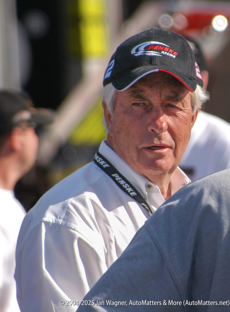 Older man wearing a Penske Racing cap and white shirt stands outdoors, looking slightly to the side. A crowd and blurred background are visible.