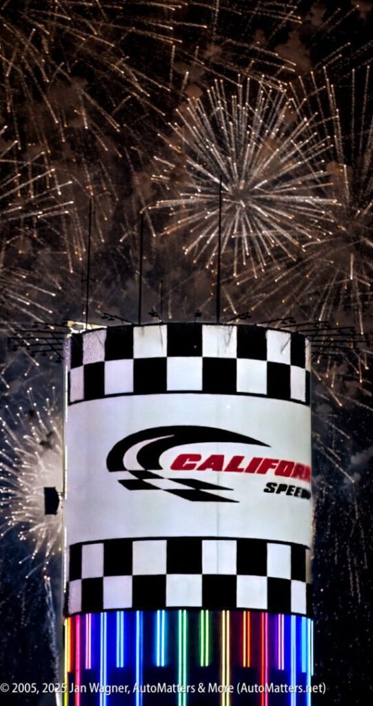 California Speedway sign with checkered flag pattern in foreground, fireworks exploding in the night sky above, and vertical neon lights below.