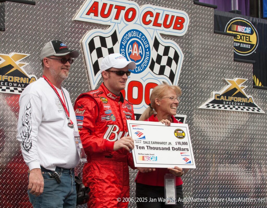 Three people stand together at an Auto Club Speedway event, with one holding a $10,000 check made out to Dale Earnhardt Jr. Racing and sponsor logos are visible in the background.