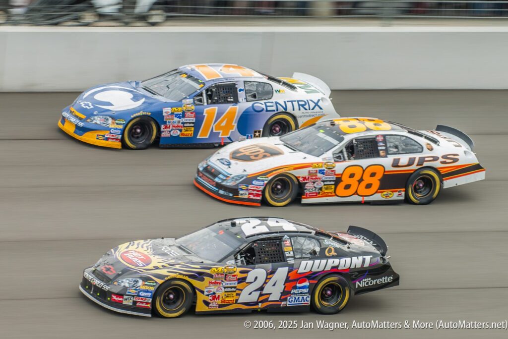 Three stock cars race side by side on a track, each with distinct sponsor logos and numbers: 14, 88, and 24. Spectators watch from behind the barrier.