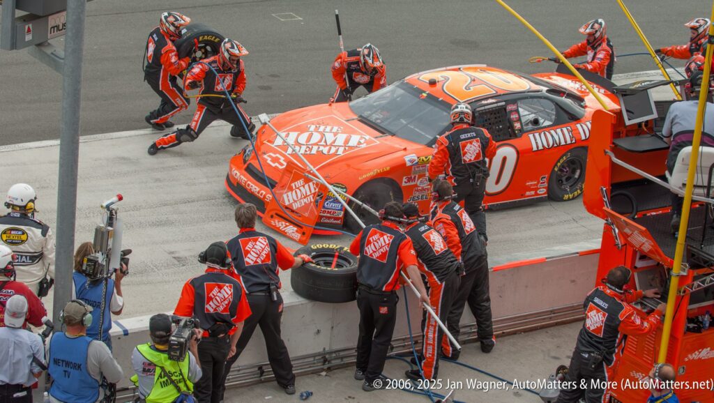A NASCAR pit crew in matching uniforms services an orange Home Depot race car during a pit stop, with crew members refueling and changing tires.