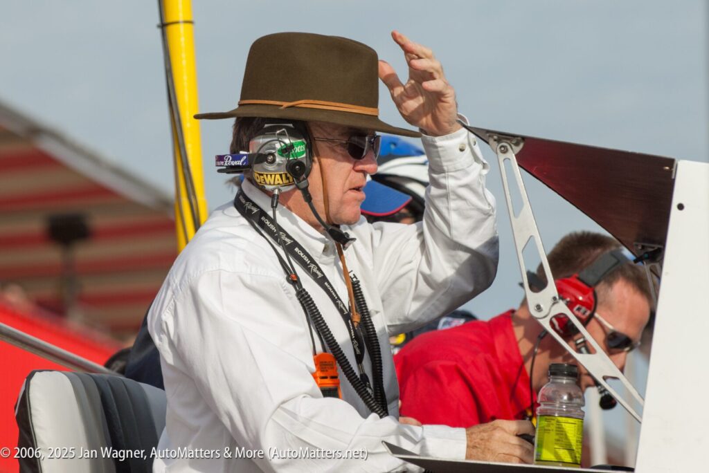A person wearing a headset and brown hat, dressed in a white shirt, stands near racing equipment, holding their hat with one hand.
