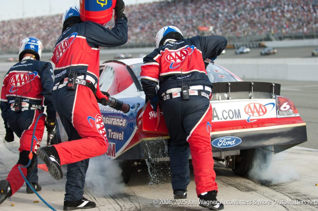 A pit crew services a Ford race car sponsored by AAA during a pit stop at a racetrack, with smoke visible and a large crowd in the background.