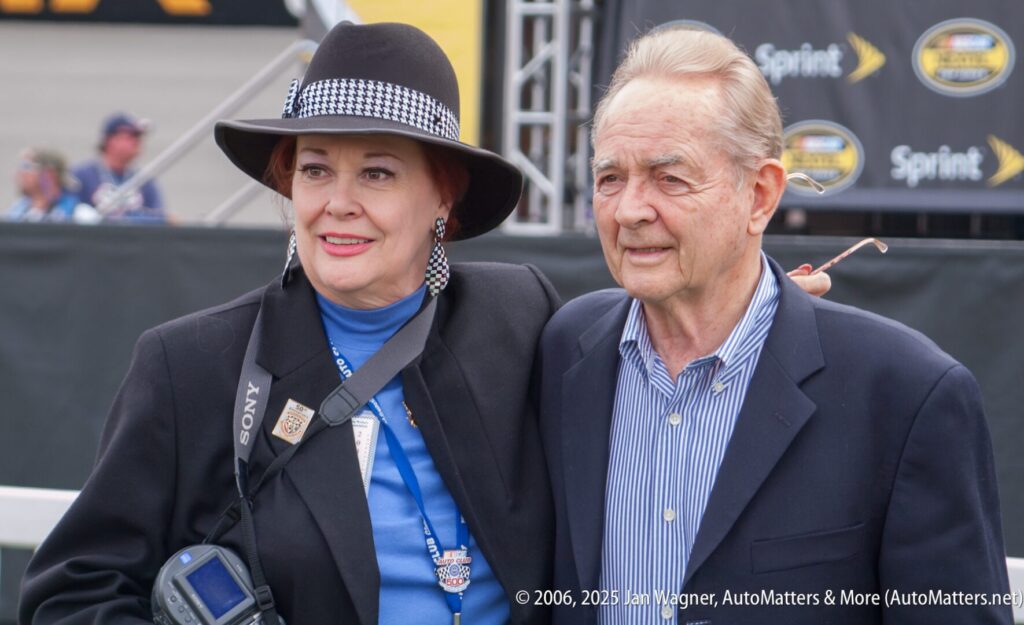 A woman in a black hat and coat stands beside an older man in a blue striped shirt and blazer at an outdoor event with Sprint branding in the background.