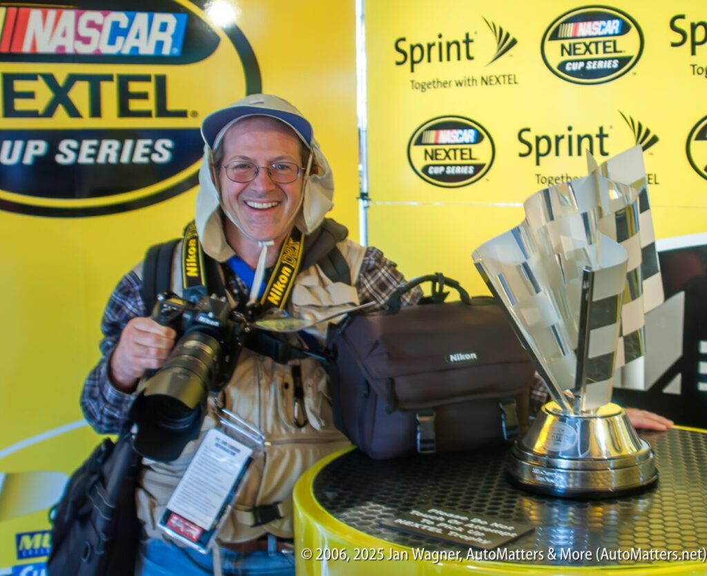 A photographer with camera gear smiles beside a large NASCAR NEXTEL Cup Series trophy on a table, in front of a branded yellow backdrop.