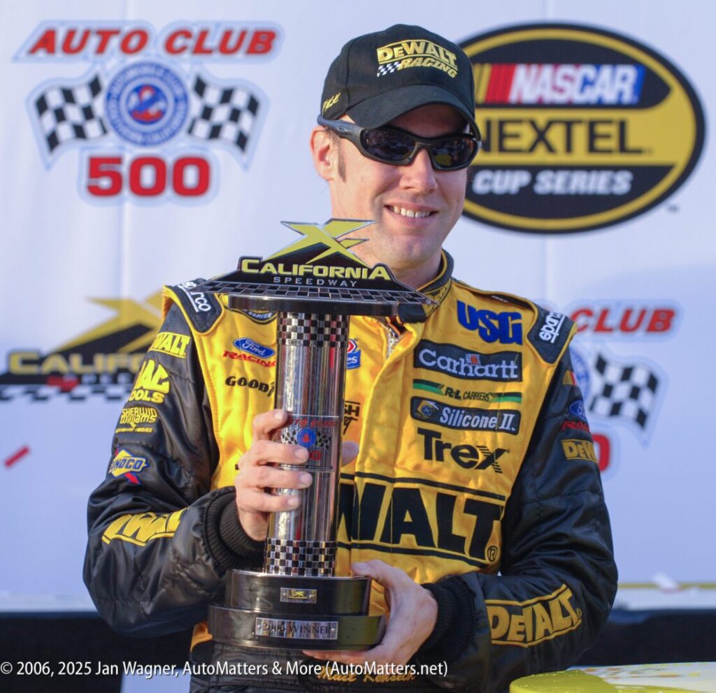 A race car driver in a yellow and black DeWalt suit and sunglasses holds a trophy in front of NASCAR and California Speedway banners.