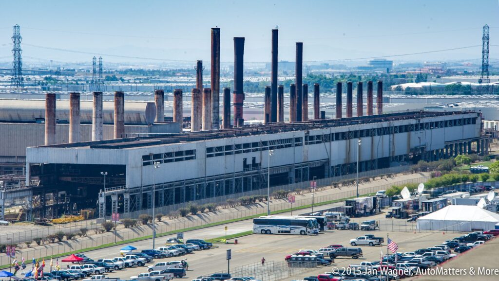 An old industrial building with numerous tall chimneys stands beside a parking lot filled with cars, buses, and tents on a clear day.