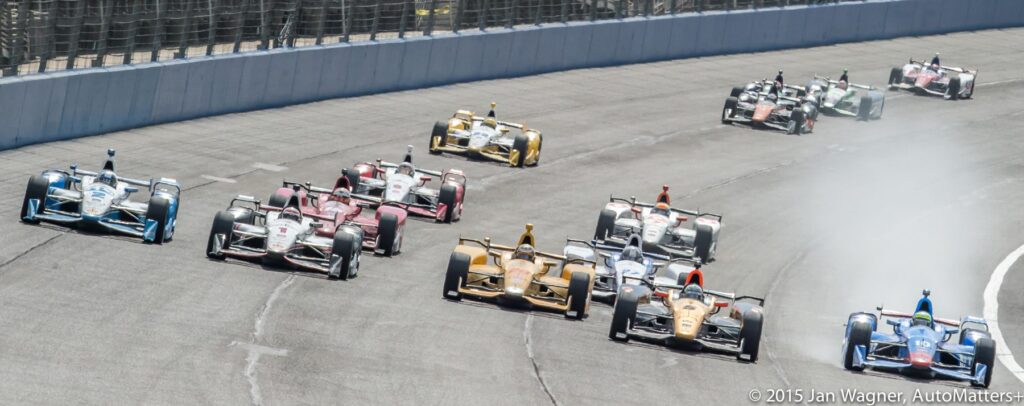 Several IndyCar race cars speed around a curved racetrack, with one car emitting smoke, during a daytime race event.