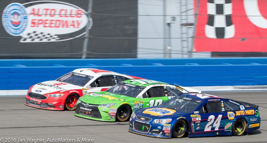 Three NASCAR race cars drive side by side on a racetrack in front of an Auto Club Speedway sign.