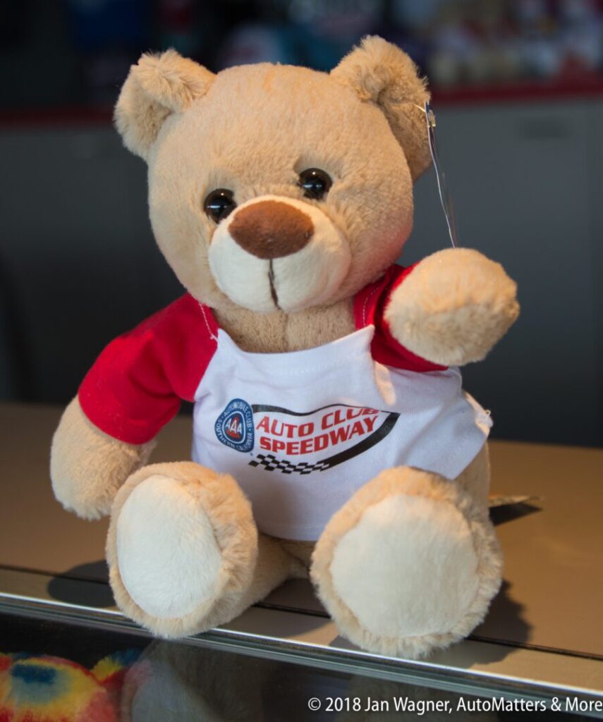 A plush teddy bear wearing a white and red Auto Club Speedway shirt sits on a reflective surface.
