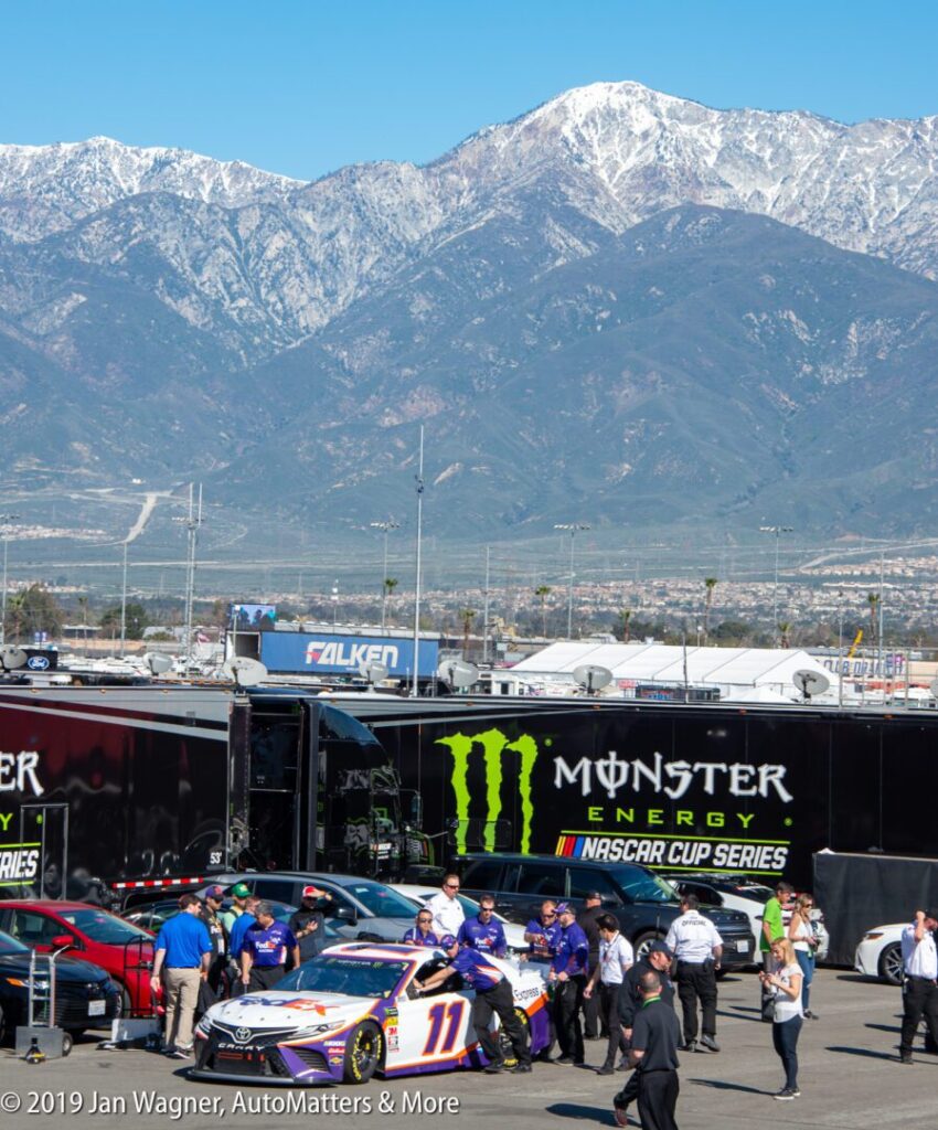 People gather around a NASCAR race car in the pit area with Monster Energy branding, set against a backdrop of mountains under a clear sky.