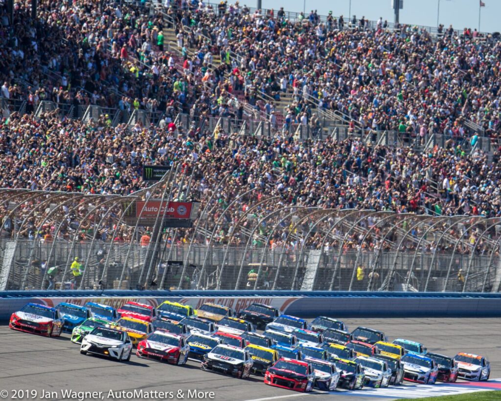 Several NASCAR race cars speed down a track side by side, with a large crowd of spectators filling the grandstands in the background.