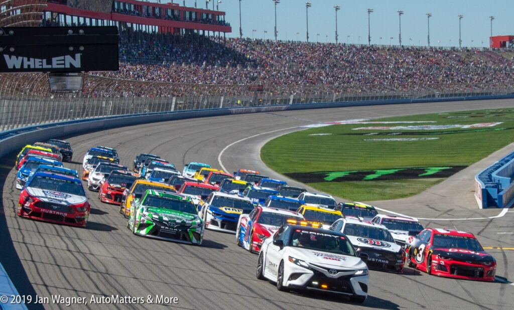 Stock cars race around a curved track at a speedway, with a large crowd in the stands and a pace car leading the pack.
