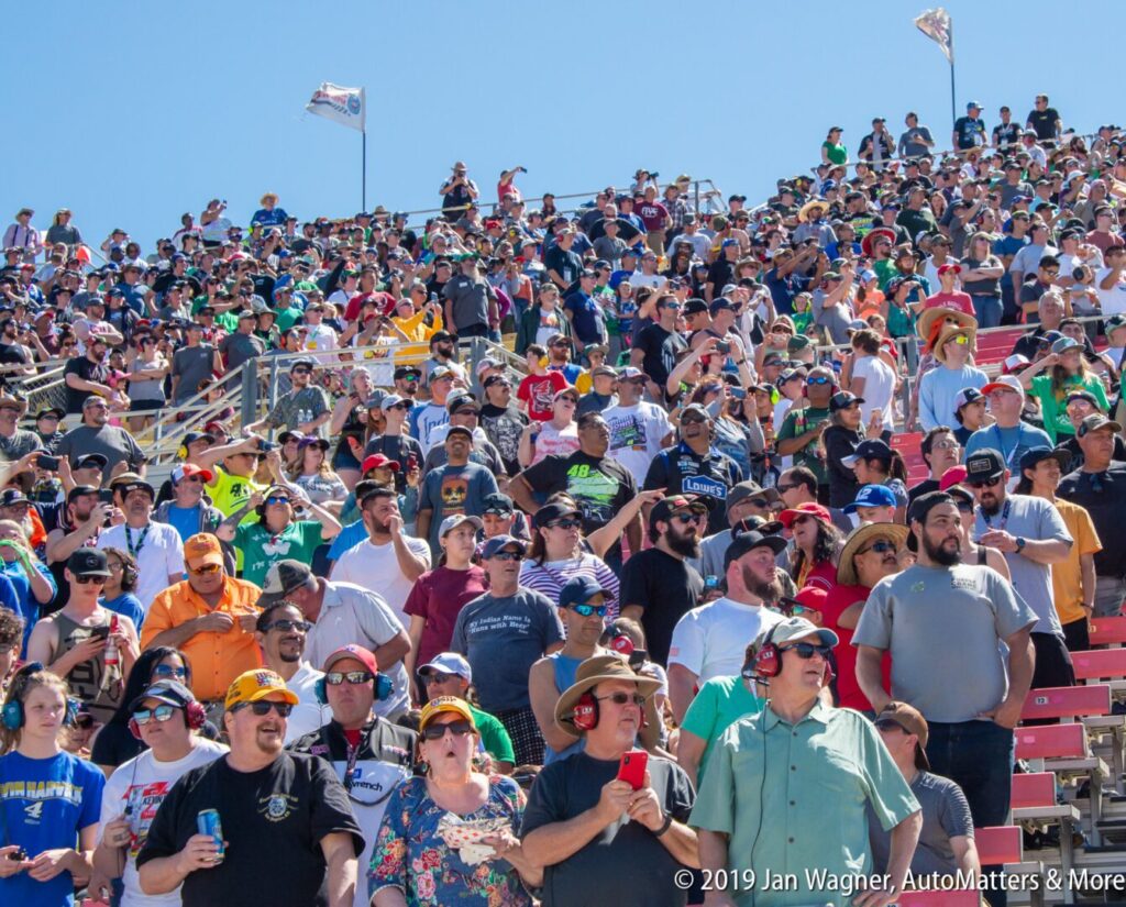 A large crowd of people fills stadium bleachers under a clear sky, many wearing hats and sunglasses, watching an outdoor event.