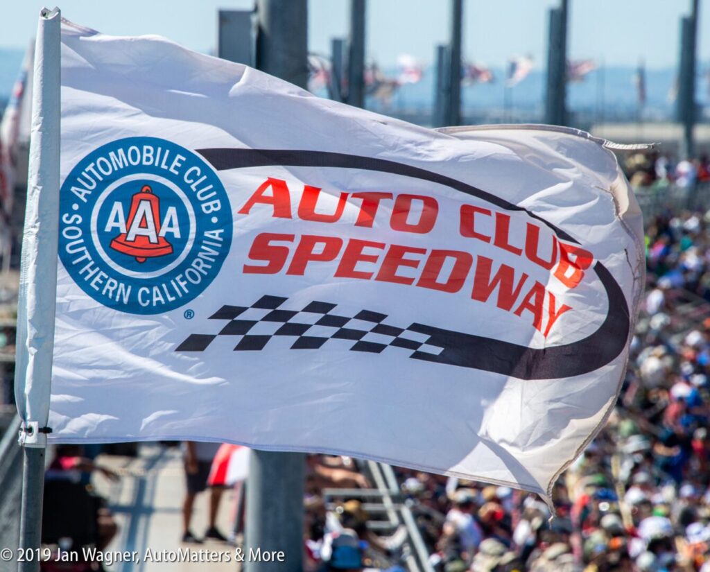 A white Auto Club Speedway flag with the AAA logo waves above a crowded grandstand at a motorsport event.