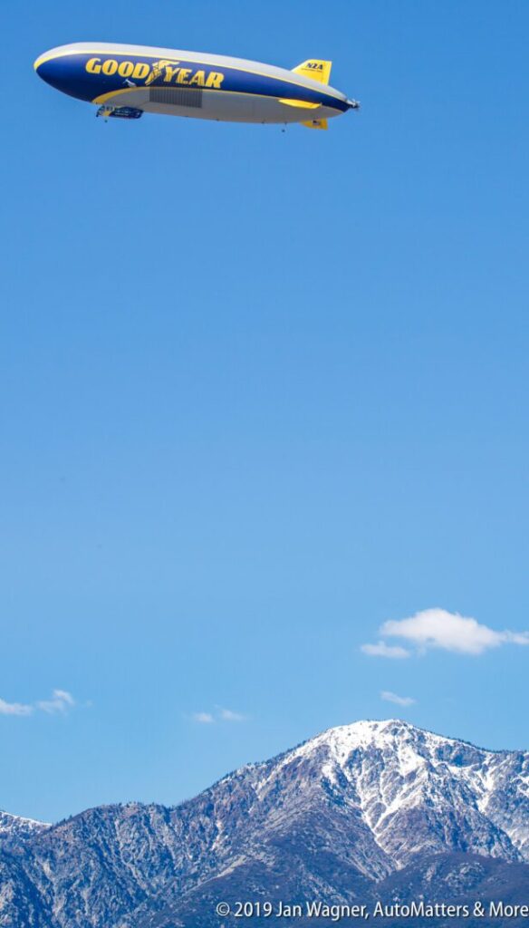 Goodyear blimp flying in a clear blue sky above snow-capped mountains.