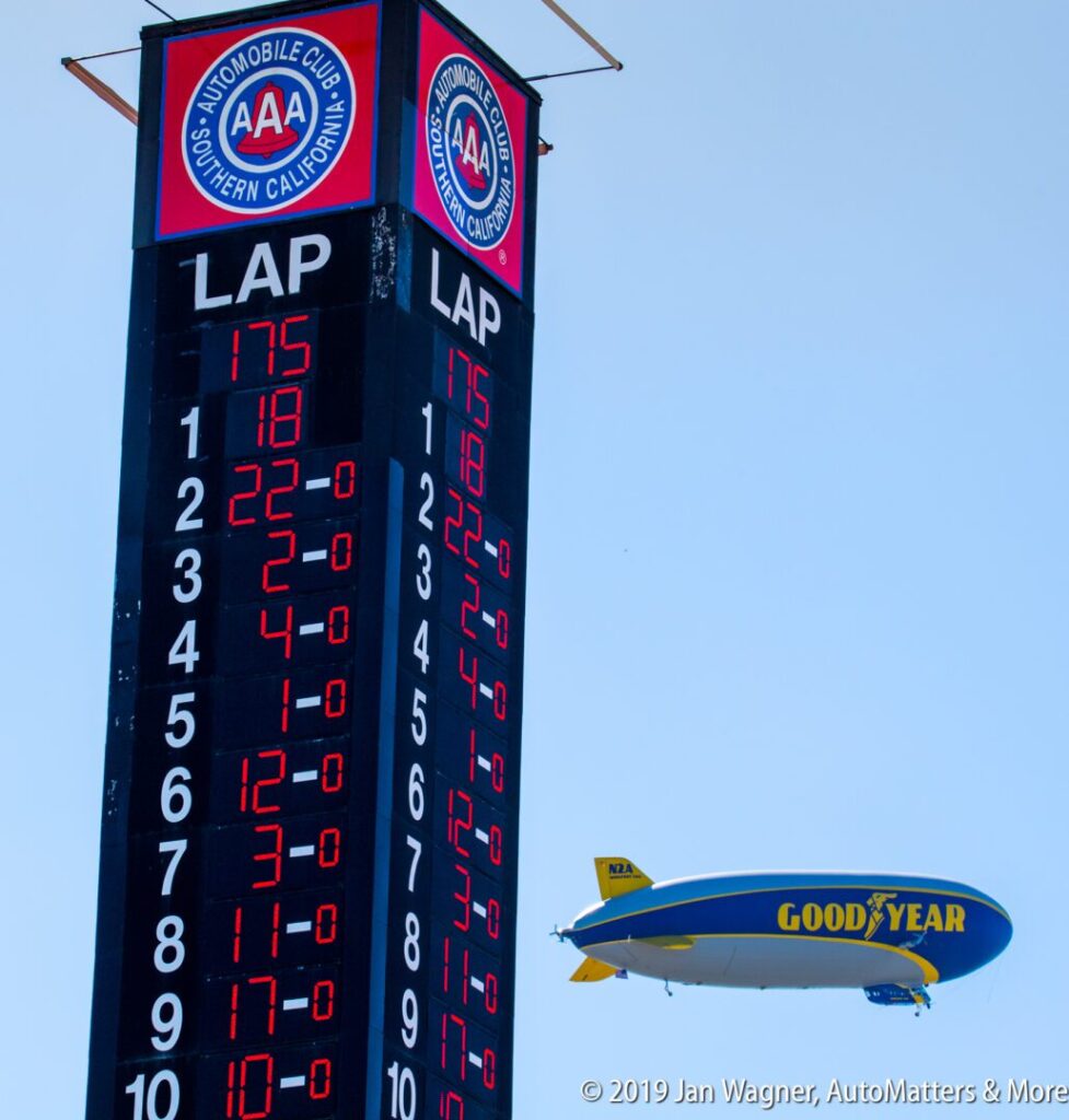 A Goodyear blimp flies near a race track scoreboard displaying lap numbers and car positions against a clear blue sky.