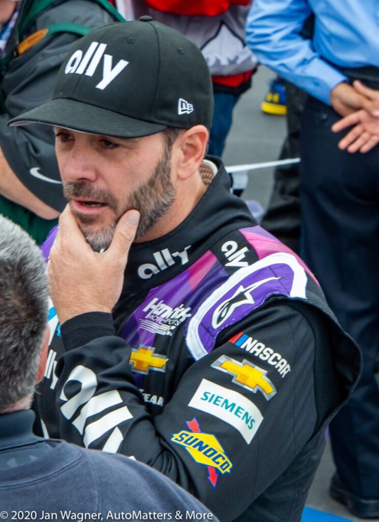A NASCAR driver in a racing suit and ally cap stands with his hand on his chin, surrounded by people. Racing sponsor logos are visible on his suit.