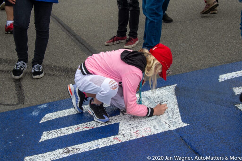 A person in a red hat and pink jacket kneels on the pavement, writing on a painted white area surrounded by others standing nearby.