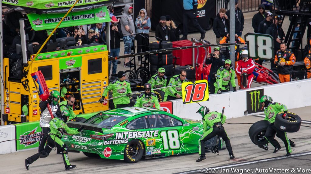 Pit crew services a green No. 18 Interstate Batteries race car, changing tires and refueling during a pit stop at a motorsport event.