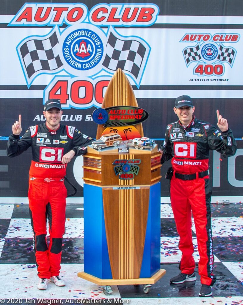 Two race car drivers in red and black uniforms pose next to a trophy at the Auto Club Speedway after the Auto Club 400 event.