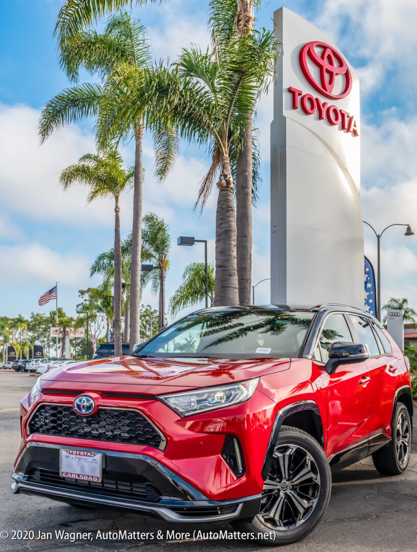 A red Toyota SUV is parked in front of a Toyota dealership sign with palm trees and flags in the background.