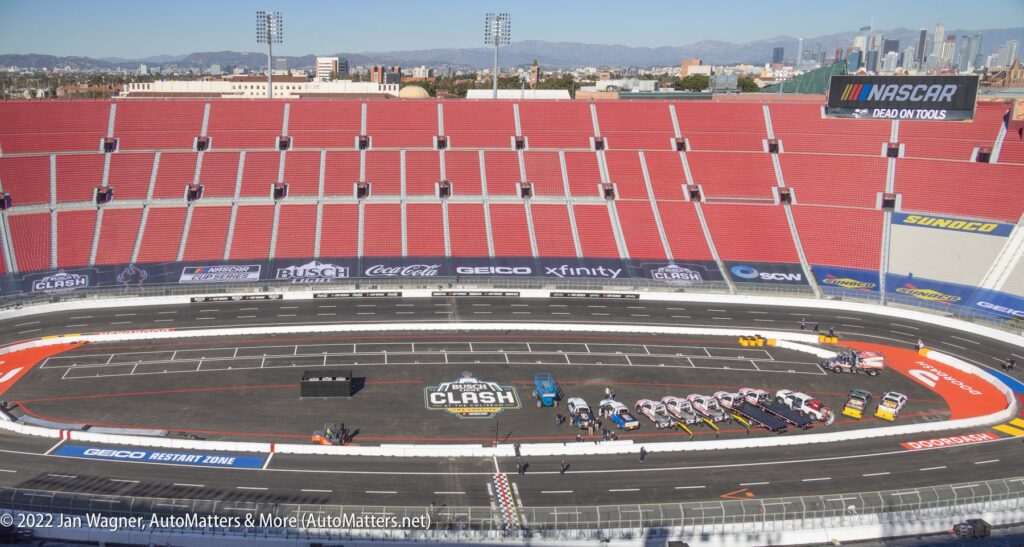 A view of the Los Angeles Memorial Coliseum set up for a NASCAR event, with race cars lined up on the track and empty red stadium seats in the background.