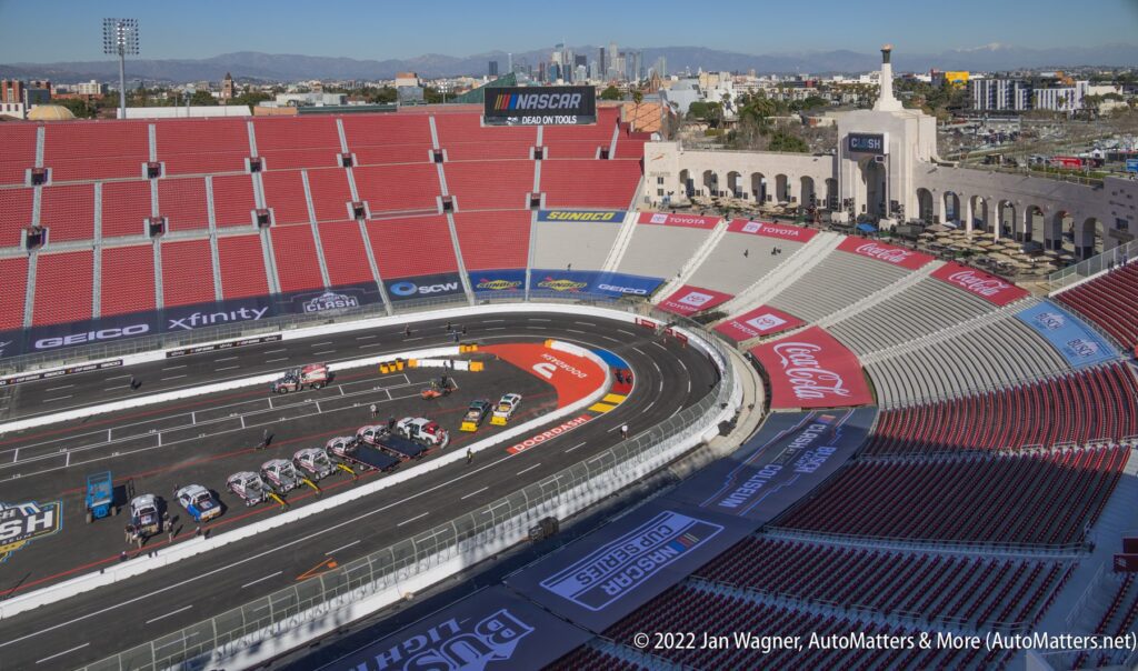 Stock cars line up on a temporary racetrack inside a large stadium with mostly empty red seats; Los Angeles skyline visible in the background.