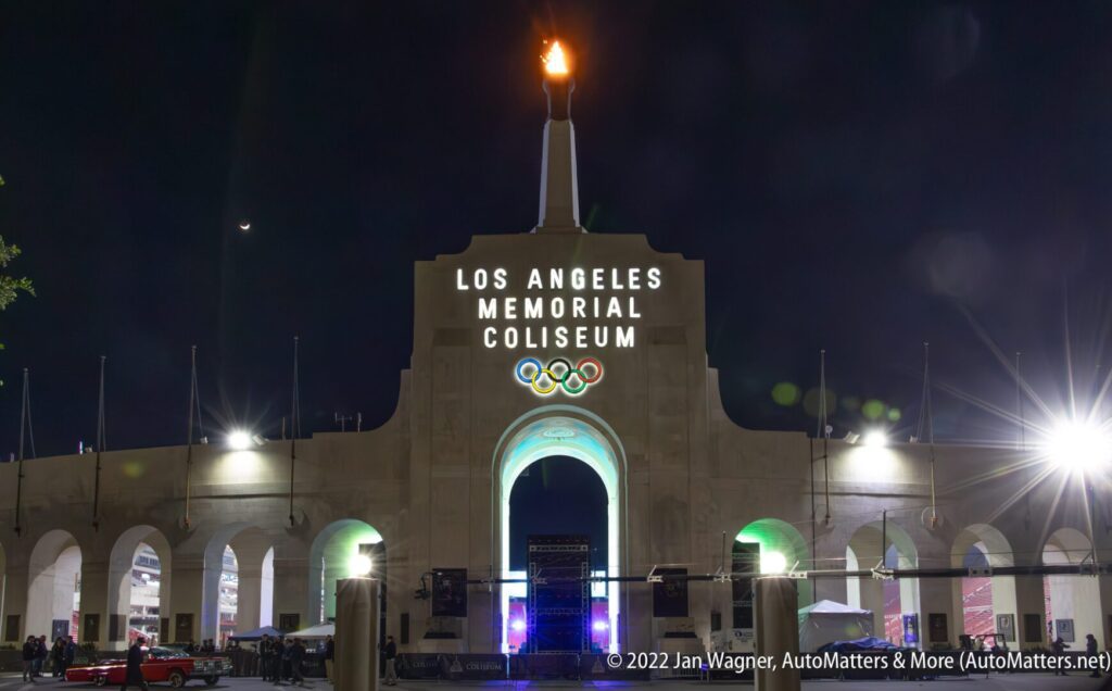 Night view of the Los Angeles Memorial Coliseum entrance, with Olympic rings displayed and a lit flame atop the structure.