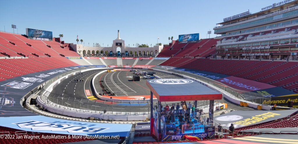 A temporary NASCAR race track set up inside a large stadium with empty red seats, broadcast booth, and sponsor banners visible.