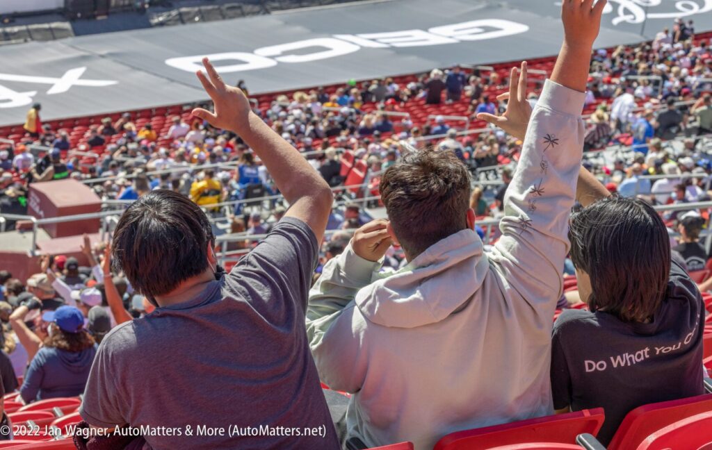Three people seated in a stadium wave their hands toward the field, where a crowd watches an event and a large GEICO logo is visible on the ground.
