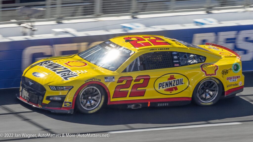 A yellow #22 Pennzoil Ford Mustang race car with Shell branding, driven by Joey Logano, on a racetrack.