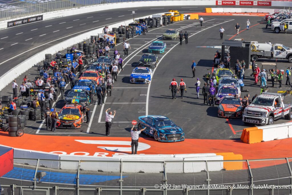 NASCAR race cars and teams prepare on the pit road before a race, with pit crew members, officials, and equipment visible around the vehicles.