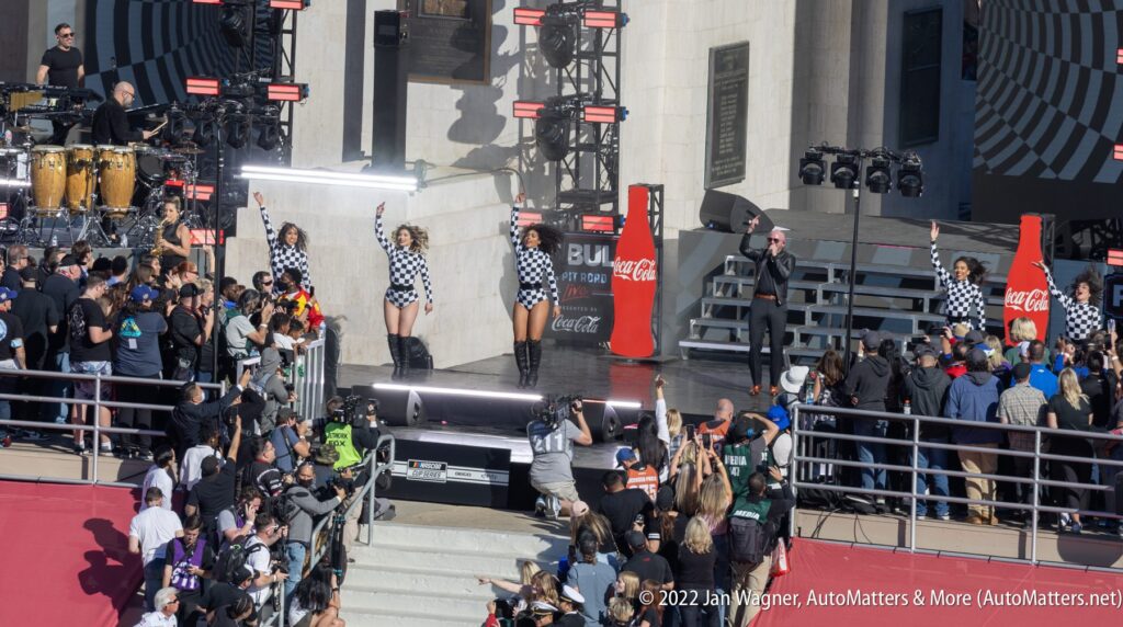 Performers in checkered outfits and boots dance on an outdoor stage as a crowd watches; large Coca-Cola bottles are visible on stage.