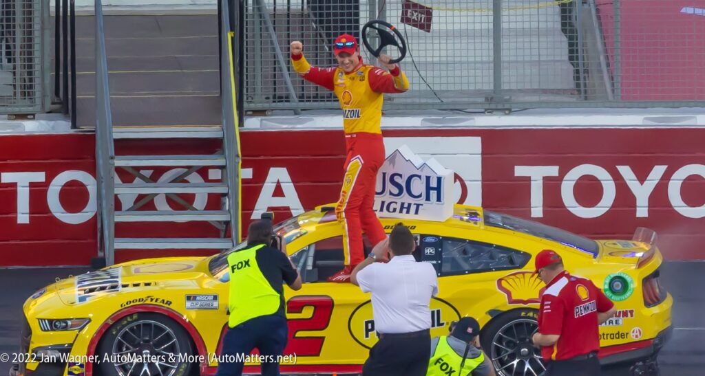 A race car driver in a yellow and red suit stands on top of a yellow car, holding a steering wheel in one hand and pointing skyward as crew members and media surround the car.
