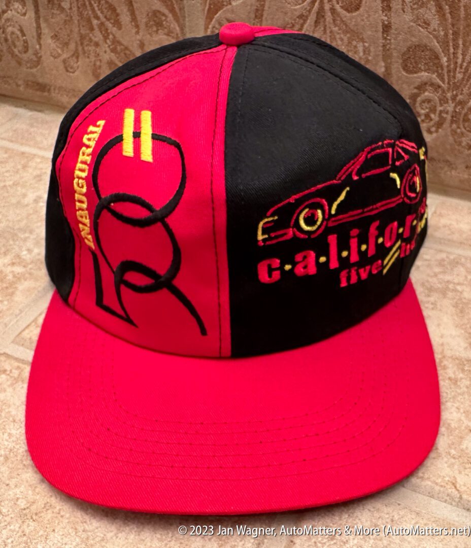Red and black baseball cap featuring embroidered graphics of a race car, the word "California", and the phrase "Inaugural five hr" on the front panels.