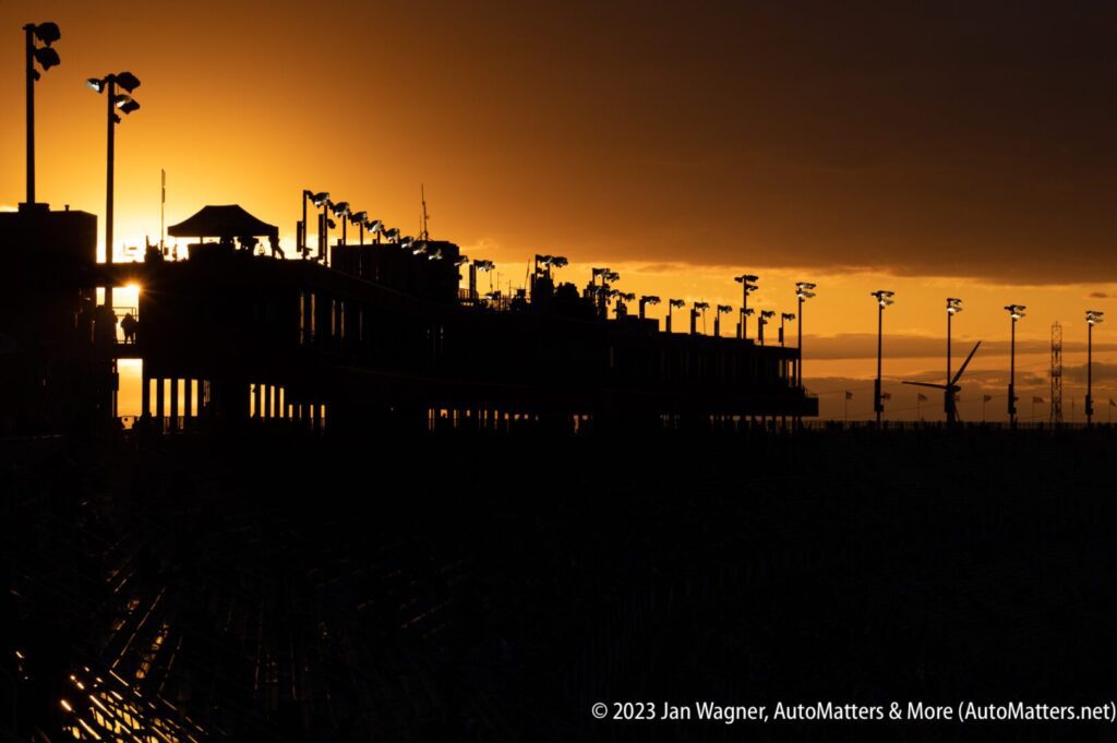 Silhouetted grandstands and light poles at sunset during an outdoor event, with a glowing orange sky in the background.