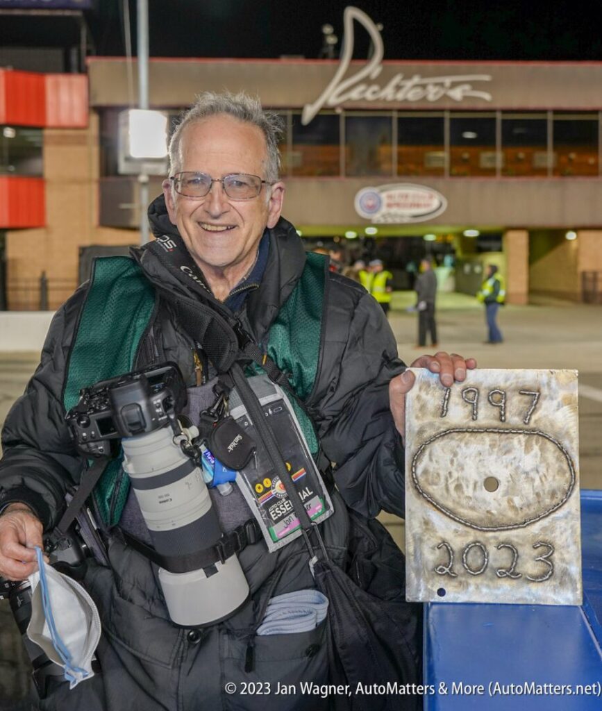 A person wearing a camera and press badge smiles while holding a rectangular plaque inscribed with "1997 - 2023" at a nighttime event outside a building.
