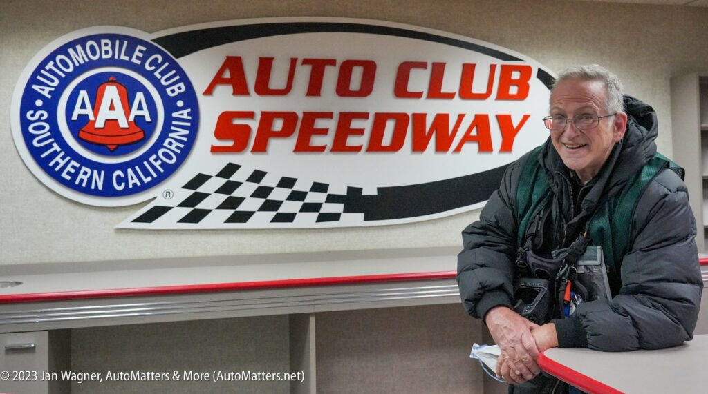 A man stands and smiles next to a counter in front of a large "Auto Club Speedway" sign at the AAA Automobile Club of Southern California.