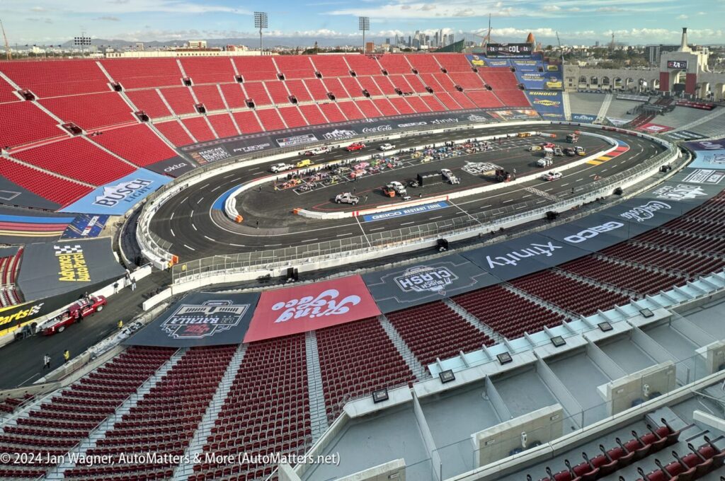 View of a large stadium with a black oval racetrack on the field, surrounded by mostly empty red seats and various sponsor banners on the ground.