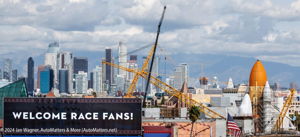 A billboard reading "WELCOME RACE FANS!" stands in front of construction cranes and buildings with the Los Angeles skyline and mountains in the background.