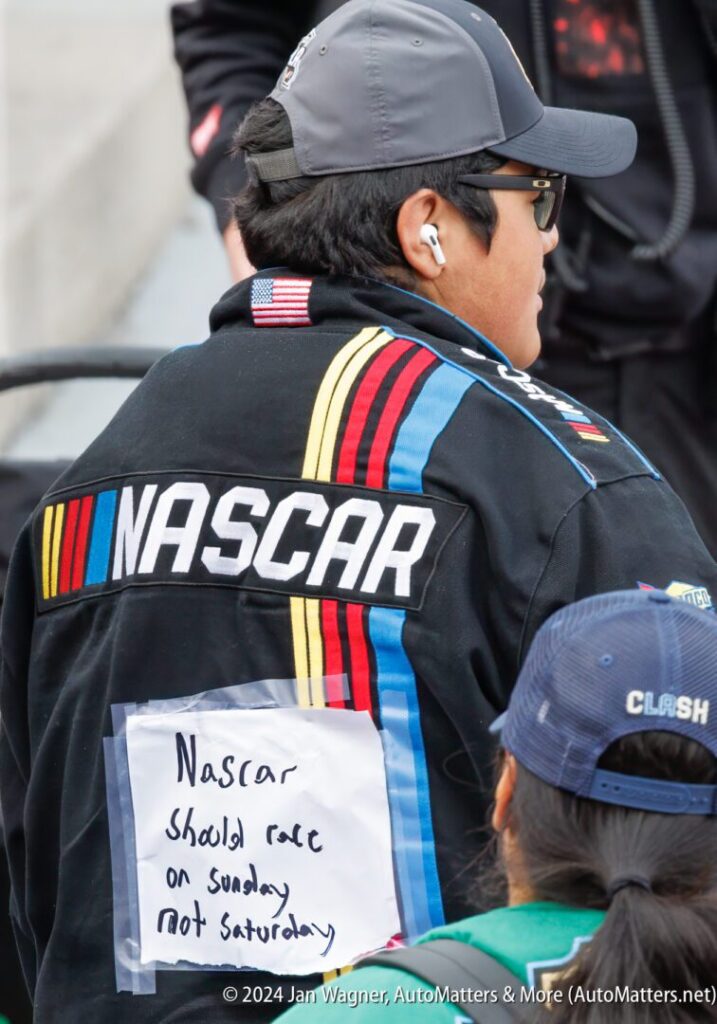 Person wearing a NASCAR jacket with a handwritten sign on the back reading, "Nascar should race on Sunday not Saturday," standing among others at an outdoor event.
