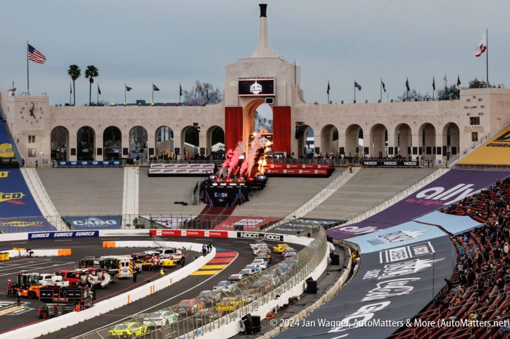 A NASCAR race takes place at a stadium with cars on the track, pyrotechnics in the background, and a large archway structure behind the stands.
