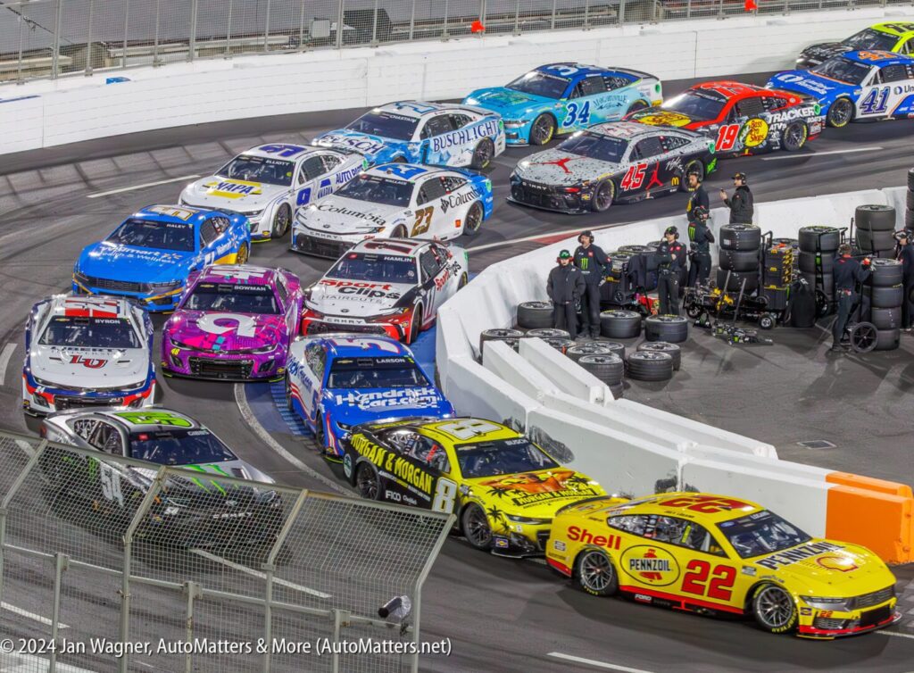 Several NASCAR race cars are bunched up on a track near a barrier, with crew members and pit equipment visible in the background.