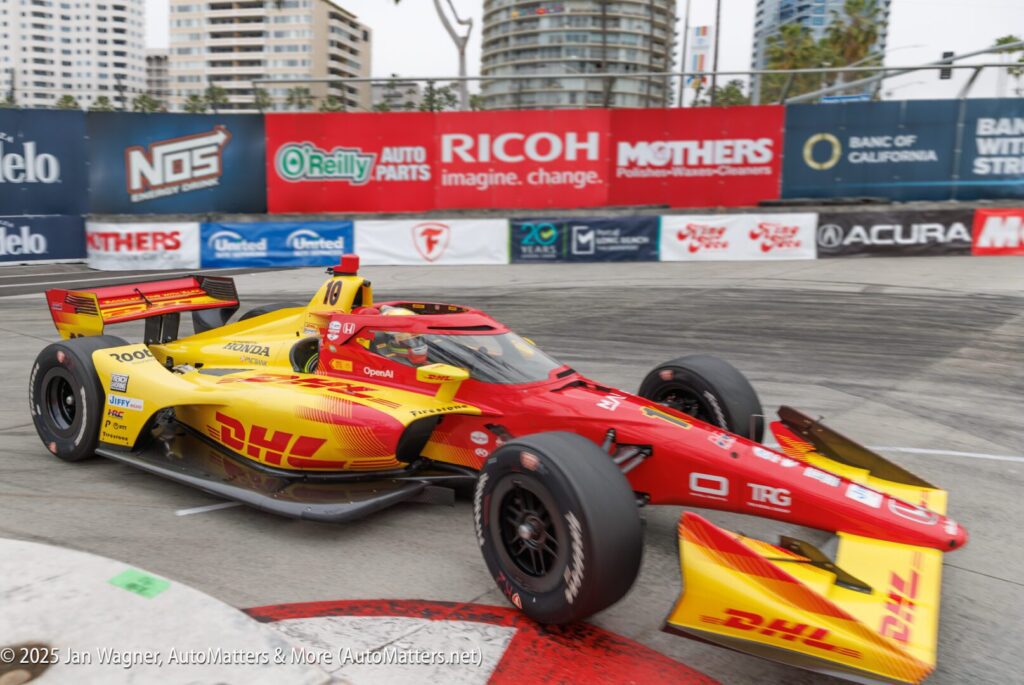 A red and yellow IndyCar with DHL branding rounds a corner on a city street circuit, passing advertising banners and barriers.