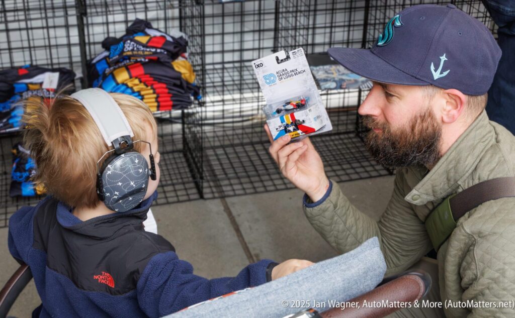 A man shows a young child wearing earmuffs a toy car package at an outdoor market stall with clothes in the background.